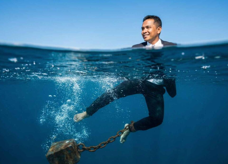 A split-level underwater photograph. Above the surface, a professional in a business shirt smiles calmly under a blue sky. Below the surface, their legs kick frantically against a heavy, rusted chained weight pulling them down into the deep blue water.