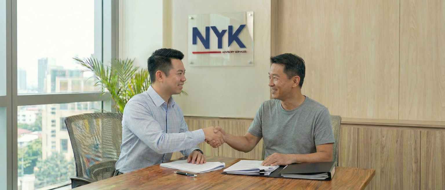 Eric smiling warmly while shaking hands with a middle-aged Chinese male client wearing a t-shirt across a wooden desk in a bright, modern office setting.