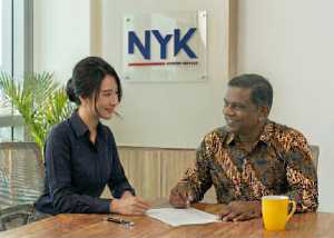 A female financial advisor at NYK Advisory Services explaining documents to a smiling male client wearing a traditional batik shirt during a consultation.